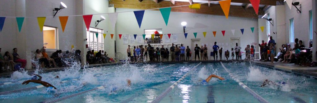 6 swimmers are coming towards you. They are each in their own lane and are racing in a swim meet. Around the pool there are people sitting or standing and watching the race.