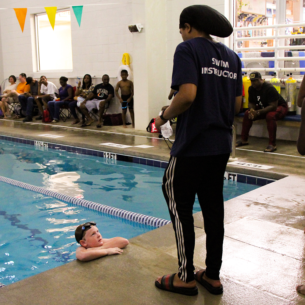 A swim instructor is talking with a young swim team member after he finishes his race. He is still in the water, using his arms on the deck to keep himself on top of the water. Around them, parent and spectators are seated to watch the race.