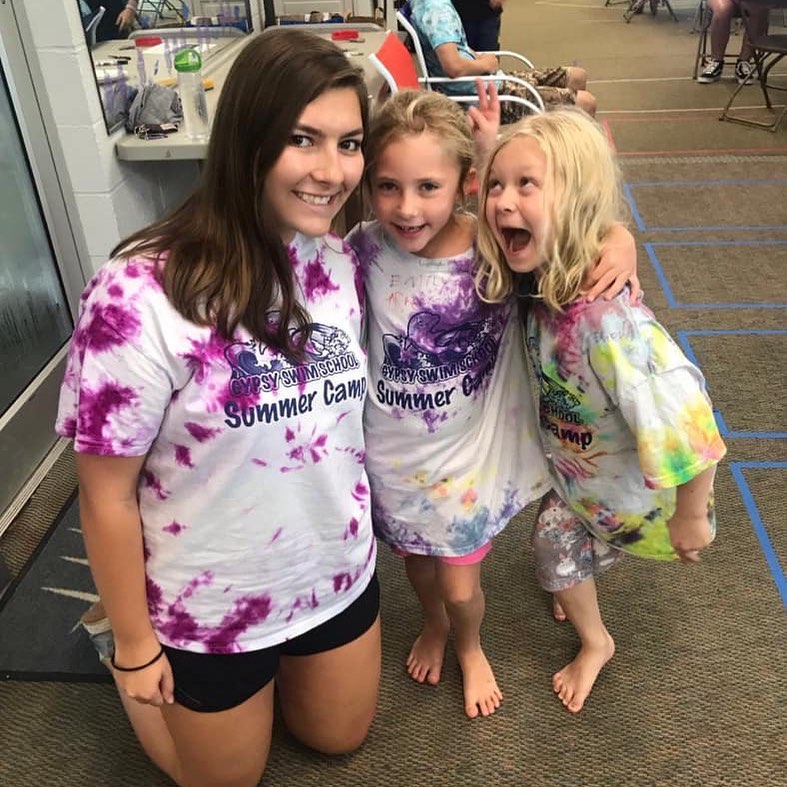 A camp staff member is kneeling on her knees with two campers beside them. All three are wearing tie dyed shirts and smiling at the camera.