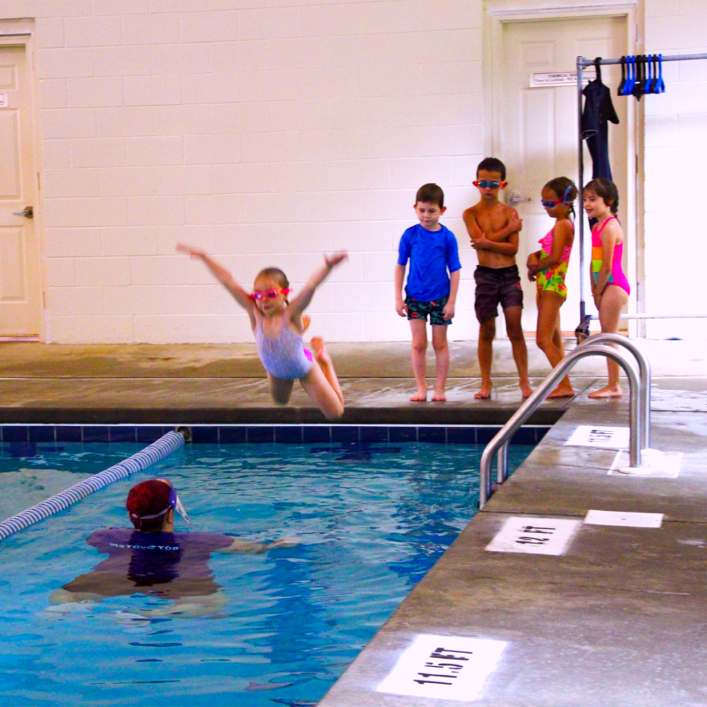 Four students are standing in line on the deck behind the deep end of the pool. One student is jumping into the pool and is midair with her arms and legs spread out wide. Below, an instructor waits in the deep end to support the student if the student needs it.