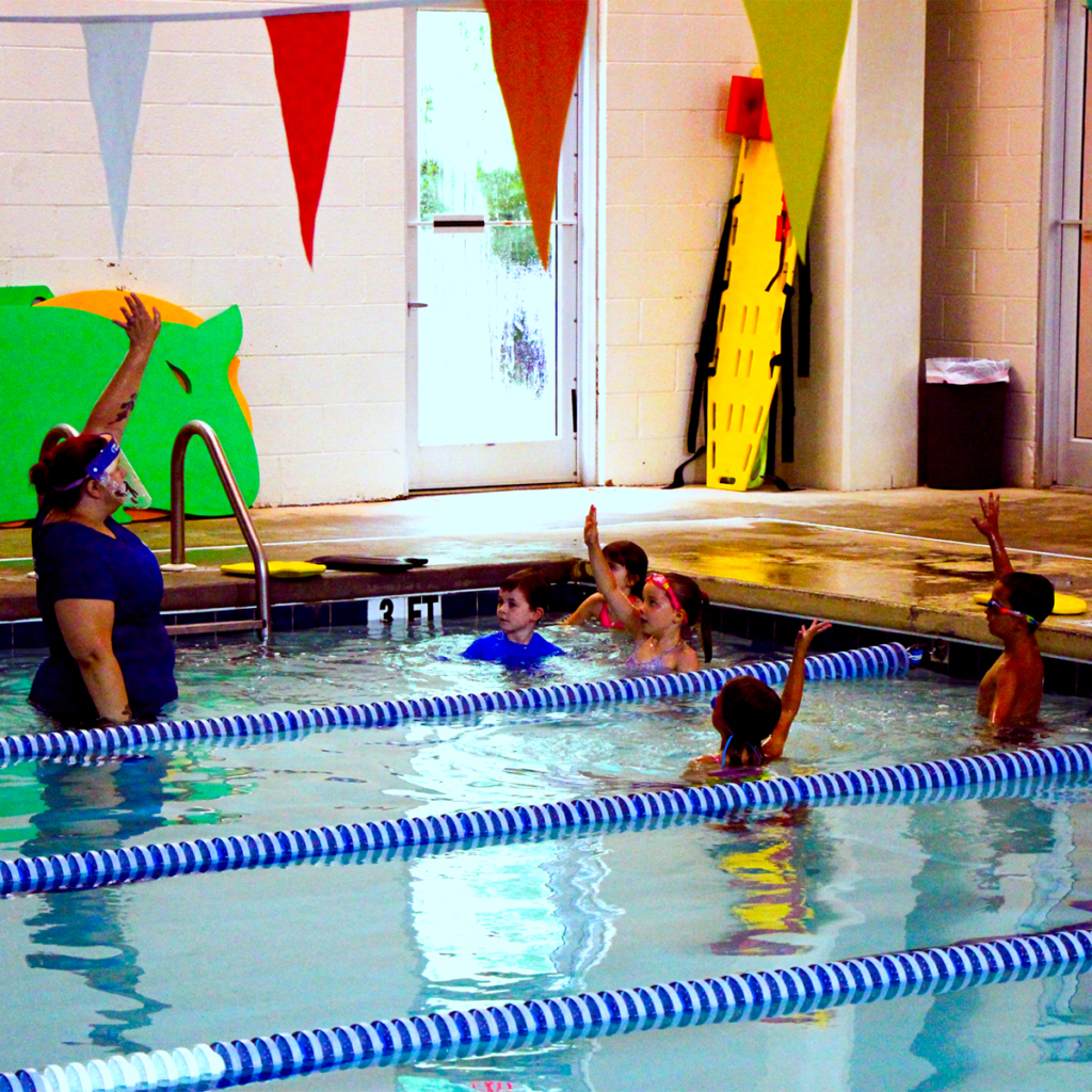 Five students stand in the shallow end of the pool. An instructor is in front of them and holding her arm in the air. Three of the five students are following her lead and are also raising their arm.