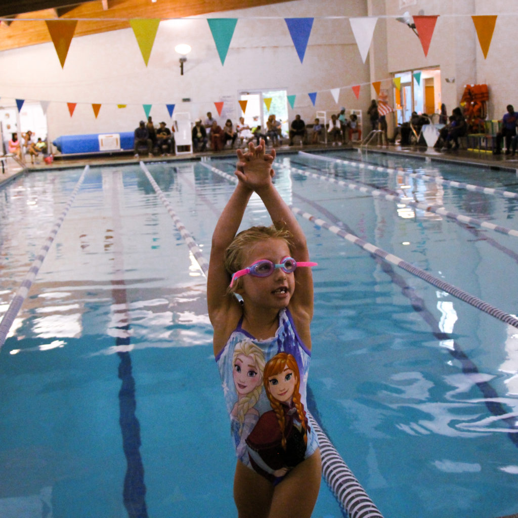 A swimmer stands before the deep end of the pool. She is turned around looking behind with her arms held above her head in a dive form.