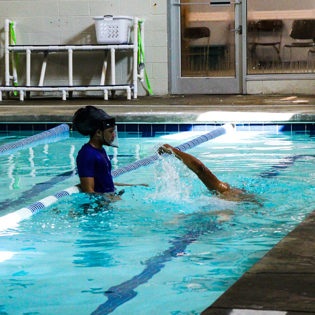 An adult swim student is practicing his freestyle stroke while his swim instructor watches.
